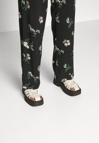 Białe sandały na cienkich paskach z grubą czarną podeszwą, noszone z czarnymi spodniami w kwiatowy wzór z różowymi i zielonymi akcentami.