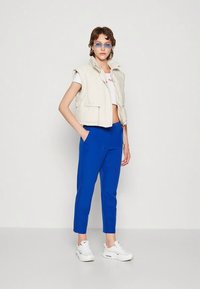 White cropped padded vest, white graphic t-shirt, bright blue tapered pants, and white sneakers. Clear sunglasses complete the outfit.