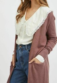 Lång vinröd kardigan med ribbade manschetter och fickor, lagd över en vit skjorta med en stor krage kantad av spets. Blå jeans synliga.