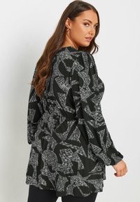 Robe noire avec un motif abstrait blanc, manches longues, taille cintrée et coupe loose. Tissu lisse avec un léger éclat.