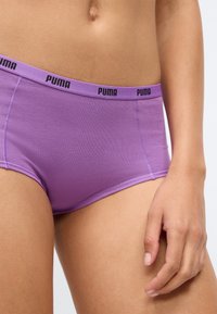 Fioletowe bawełniane slipy z czarnym logo "PUMA" na pasku, z bezszwowym designem i gładką teksturą. Ręka oparta na udzie.