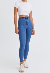 Jean en denim bleu taille haute avec une coupe slim, présentant un design classique à cinq poches et une texture délavée. Associé à des baskets blanches.