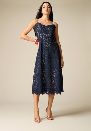Frau trägt ein marineblaues Spitzen-Midikleid mit dünnen Trägern und gewelltem Saum, kombiniert mit roten Armreifen und Riemchenabsätzen.