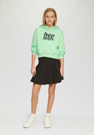 Miętowa bluza z kapturem z grafiką "bądź miły", zestawiona z czarną plisowaną spódnicą. Modelka nosi białe sneakersy z falbanym skarpetami.
