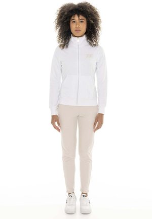 Une femme se tient face à l’objectif, vêtue d’une veste blanche zippée, d’un pantalon beige et de baskets blanches, sur un fond uni.