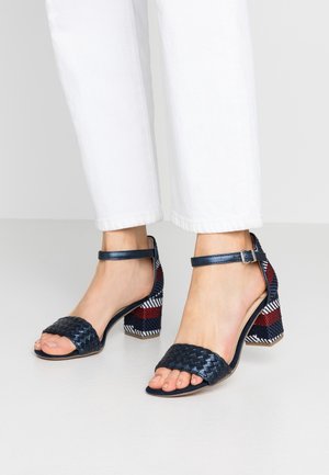 Pies calzados con zapatos de tacón abiertos de cuero trenzado azul marino, con tiras en el tobillo y tacones anchos que presentan patrones a rayas en rojo, blanco y azul.
