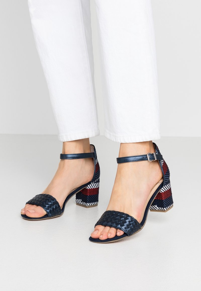 Pieds portant des sandales à talons ouverts en cuir tressé bleu marine avec brides à la cheville et talons blocs ornés de motifs rayés rouge, blanc et bleu.