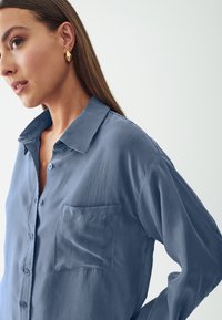 Blusa in cotone azzurro chiaro con abbottonatura frontale, colletto a punta e taschino sul petto. Presenta maniche lunghe e una texture morbida e liscia.