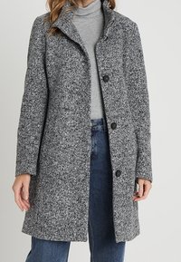 Manteau gris texturé avec un col châle et trois boutons noirs. Porté par-dessus un col roulé gris et un jean bleu.