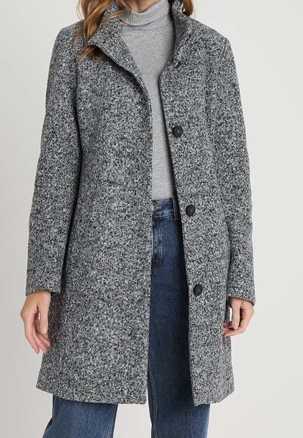 Manteau gris texturé avec un col châle et trois boutons noirs. Porté par-dessus un col roulé gris et un jean bleu.