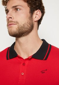 Rotes Poloshirt mit schwarzem Kragen, ausgestattet mit zwei Knöpfen und kontrastierenden Nähten. Gesticktes Logo auf der linken Brust. Baumwollmaterial.