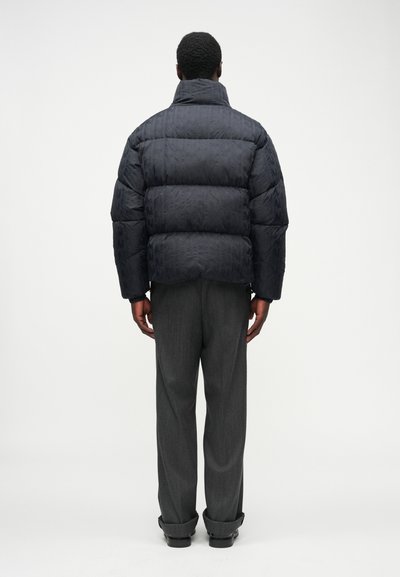 Jachetă puffer de culoare bleumarin închis, cu un design matlasat și guler înalt. Asortată cu pantaloni gri, evidențiind o croială relaxată și mansete răsucite.