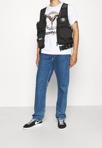 Chaleco de utilidad negro con múltiples bolsillos, llevado sobre una camiseta blanca con diseño gráfico. Combinado con vaqueros azul claro y zapatillas negras.