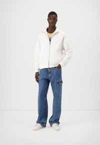 Hvit zip-up hettegenser, blå vide jeans med sidelommer, og hvite sneakers. Glatt tekstur på toppen, denim-finish på bunnen.