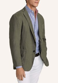 Mann trägt olivgrünen Leinenblazer über blau-weiß gestreiftem Hemd mit rotem Innenkragen, kombiniert mit weißer Hose und braunem Gürtel.