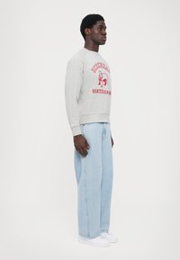 Grå sweatshirt med rød grafisk tekst og billede, parret med lyseblå baggy jeans og hvide sneakers, vist i profil.