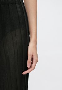 Fekete bordázott hosszú ruha áttetsző anyaggal, függőleges csíkokkal és elasztikus derékszorítóval. Egy kéz nyúlik ki az oldalról.