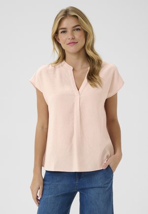 Blouse rose clair à manches courtes, avec un décolleté en V partiel et une coupe décontractée. Texture lisse et ourlet droit, associée à un jean bleu foncé.