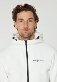 Chaqueta blanca con capucha, cierre frontal y cordón, con detalles en negro y el logo "SAIL RACING" en el lado izquierdo del pecho.