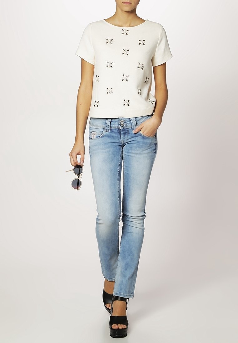 Top corto blanco de manga corta con adornos metálicos, combinado con jeans rectos de mezclilla azul claros y sandalias de plataforma negras.