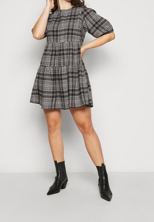 Kobieta w krótkiej sukience w czarno-białą kratę z bufiastymi rękawami i czarnych botkach, stojąca na tle jednolitego jasnego tła.