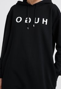 Sudadera negra con un gráfico frontal en letras blancas. Incluye capucha con cordón y puños acanalados, hecha de material suave.