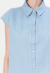 Camicia in denim azzurro chiaro con colletto classico, maniche corte e abbottonatura frontale con sei bottoni argento e una leggera trama.