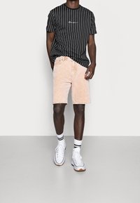 Černé pruhované tričko s bílým logem, světle růžové kordové šortky a bílé tenisky s černobílými ponožkami, stojící na neutrálním pozadí.