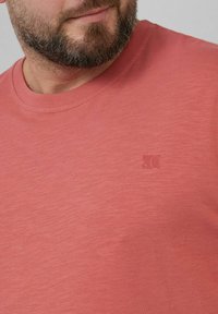 Kurzärmliges T-Shirt aus weichem Koralle-Stoff mit Rundhalsausschnitt, das ein dezentes geprägtes Logo auf der Brust und eine glatte Textur aufweist.