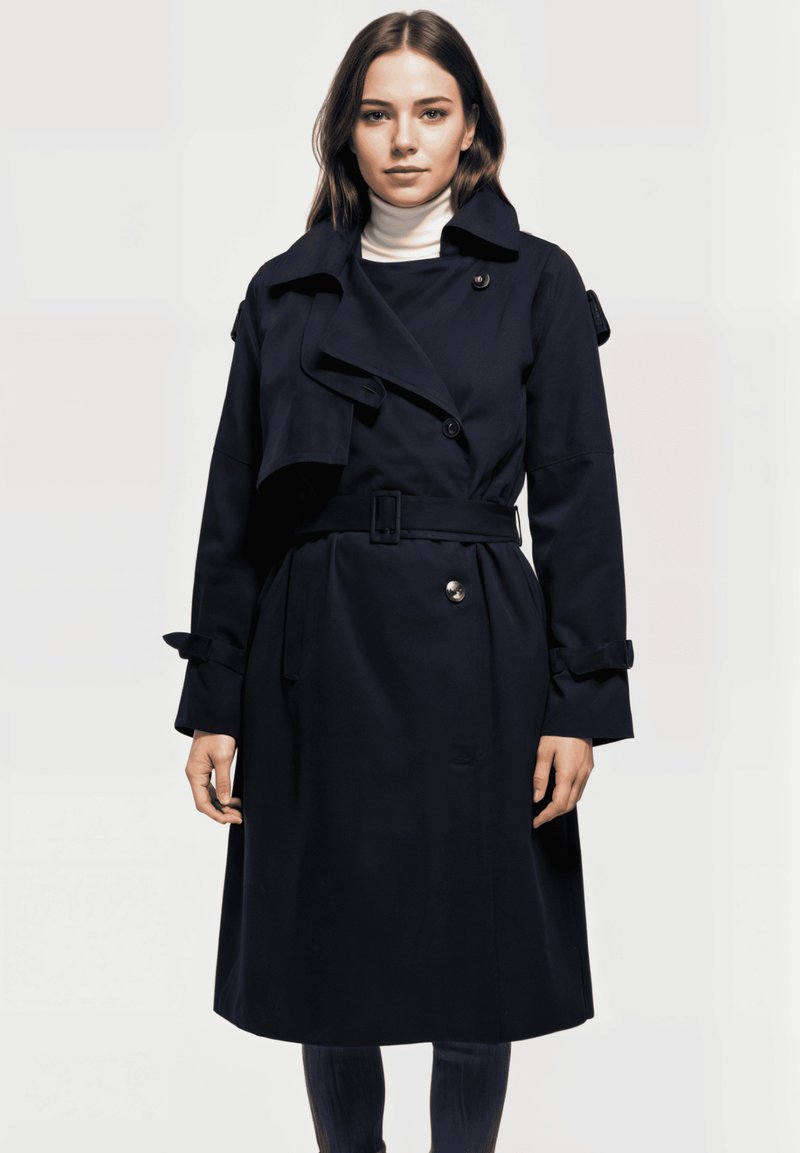 Mujer vestida con un abrigo de trench azul marino con cinturón sobre un suéter de cuello alto blanco, de pie frente a un fondo liso claro, mirando hacia adelante.