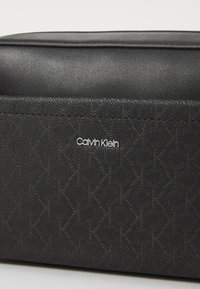 Fekete Calvin Klein táska sima és texturált anyagokból, dombornyomott geometrikus mintákkal és egy ezüst színű logóval az elején.