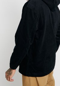 Black corduroy jacket with a hood, featuring a textured surface and elasticated cuffs, shown from a side angle. Tan pants are worn below.