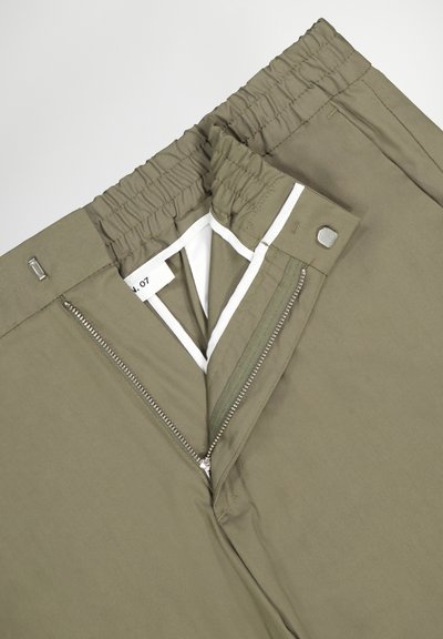 Pantalons vert olive avec une taille élastique, une fermeture éclair argentée et une doublure intérieure blanche contrastante. Présentent une texture lisse et une coupe soignée.