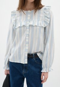 Blusa a righe blu chiaro e bianco con spalline e polsini arricciati, dotata di chiusura a bottoni sul davanti e vestibilità rilassata. Abbinata a jeans blu scuro.