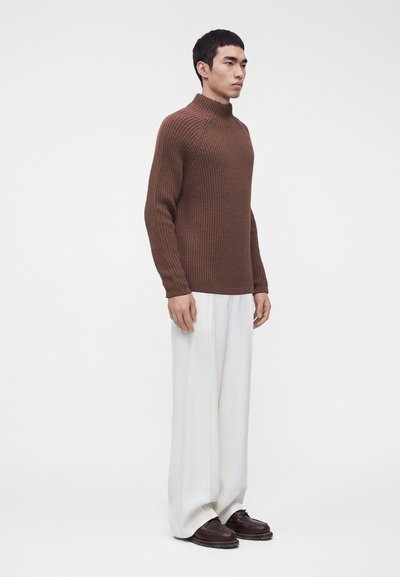Homme debout portant un pull à col roulé côtelé marron, un pantalon blanc ample et des chaussures marron foncé sur un fond blanc uni.