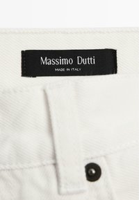Vitt tyg med texturerad yta, med en svart etikett som säger "Massimo Dutti" och "Made in Italy." Inkluderar en svart knäppknapp.
