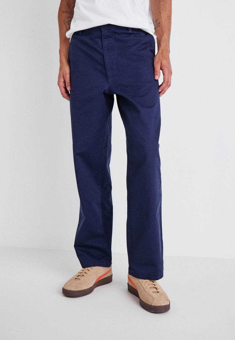 Navy blue straight-leg pants made of cotton, featuring a zip fly and two front pockets. Paired with light brown sneakers with orange accents.