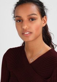 Sudraba kaklarota ar apaļu piekariņu, ko nēsā modele. Modele ģērbusies maroon krāsas ribotā topā. Maiga apgaismojums izceļ iezīmes.