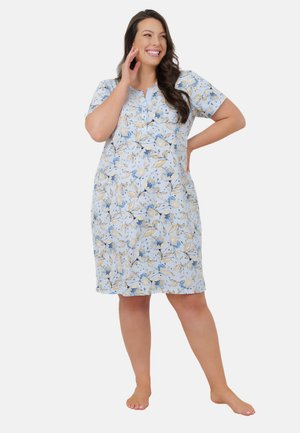 Uśmiechnięta kobieta stoi boso w sukience do kolan z krótkim rękawem, w kwiaty w odcieniach niebieskiego i beżu na białym tle.