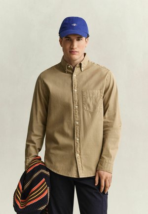 Junger Mann trägt ein beiges Hemd mit Knöpfen und eine blaue Kappe, hält einen gestreiften Pullover vor einem schlichten hellen Hintergrund.