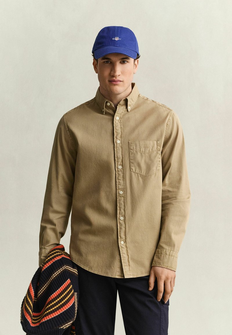 Young man wearing a beige button-up shirt and blue cap holding a striped sweater against a plain light background.