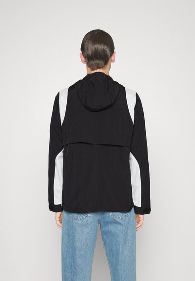 Schwarze und graue Kapuzenjacke mit lockerem Schnitt, aus strukturiertem Stoff, mit elastischen Bündchen und einem Rückenschlitz für Belüftung.