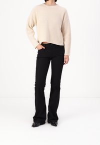 Suéter beige de tejido acanalado con un corte holgado, combinado con jeans negros acampanados. Las botas tienen puntas puntiagudas y detalles de herrajes plateados.