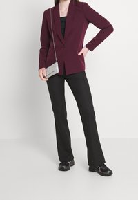 Bordeaux blazer met een sjaalkraag, twee voorzakken, gecombineerd met zwarte uitlopende broek en zwarte sneakers. Inclusief een zilveren schoudertas.