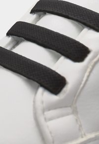 Εγγύς πλάνο λευκού αθλητικού παπουτσιού με τρία μαύρα πλεγμένα κορδόνια που περνούν από τις οπές σε επενδεδυμένο τμήμα.