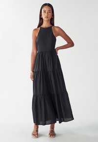 Žena oblečená v bezramínkových černých vrstvených maxi šatech a sandálech, stojící s jednou rukou na bocích před jednoduchým světlým pozadím.