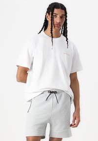 Hvit bomulls t-skjorte med korte ermer og en liten oransje logo, sammen med lysegrå shorts med snøring og sidelommer.