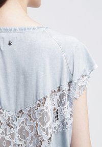 Světle modrý top s vrstveným designem a krajkovým detailem na zadní straně, krátkými rukávy a malou hvězdičkou na výstřihu.