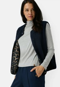 Donkerblauwe fleece vest met een voering van luipaardprint, drukknopen, gedragen over een lichtgrijs langärmige top, gecombineerd met donkere broeken.