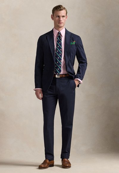 Μπλε σκούρο κοστούμι με γιακά notch, συνδυασμένο με ένα ροζ ανοιχτό ριγέ πουκάμισο και μια πράσινη γραβάτα με σχέδιο. Καφέ δερμάτινα παπούτσια.